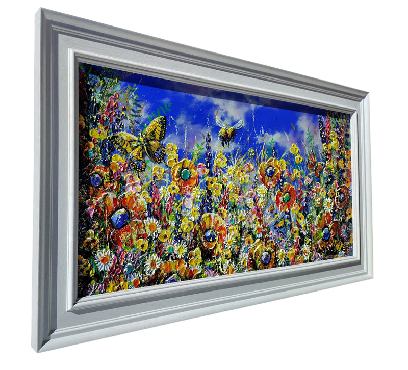 Framed wall art showing a vivid floral meadow with yellow, orange, red, and blue flowers, butterflies, and a bright blue sky, presented in a silver frame on a white background.