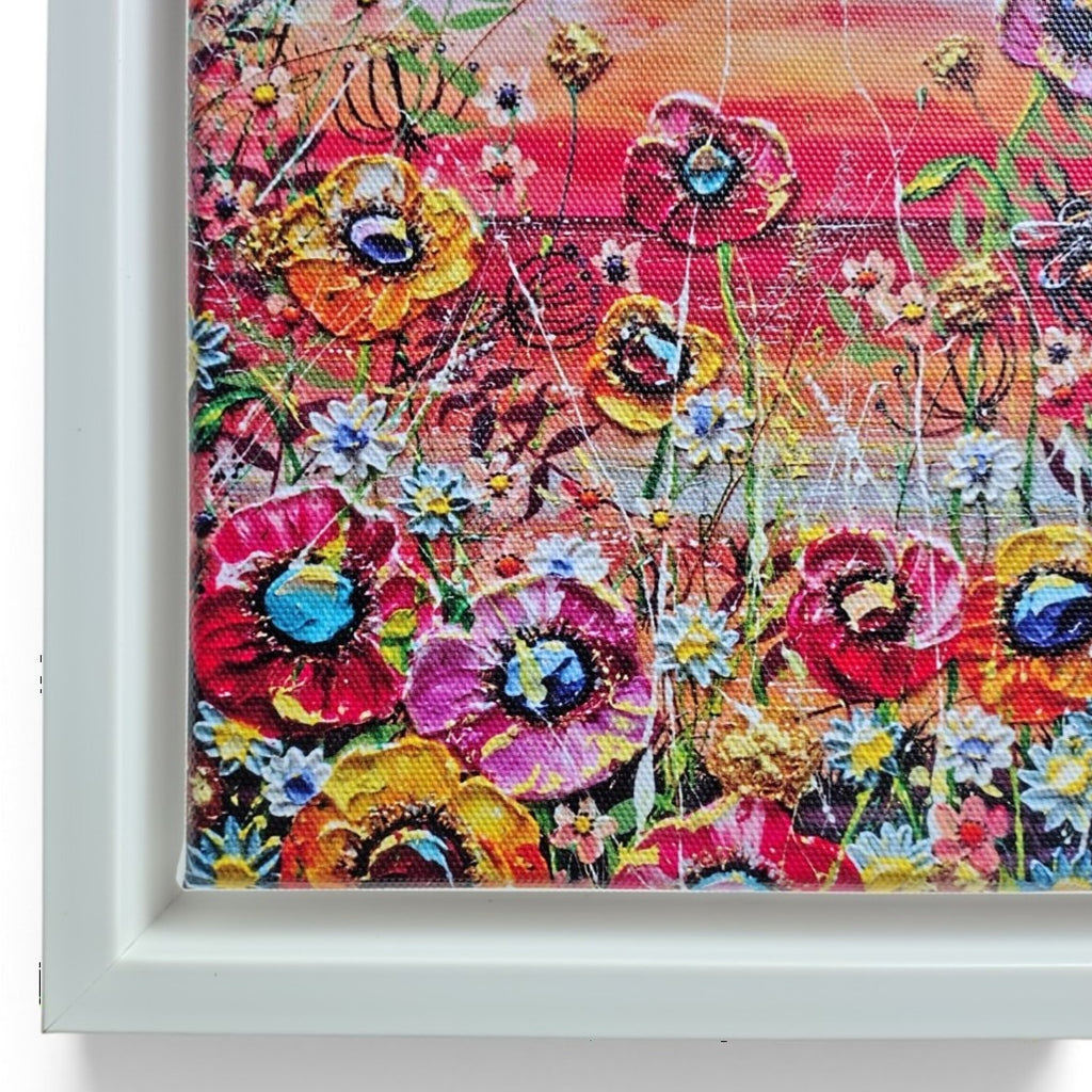 A framed floral art print featuring a vibrant, textured cross-stitch style design of poppies and wildflowers in a field under a warm orange sky, set in a white frame.