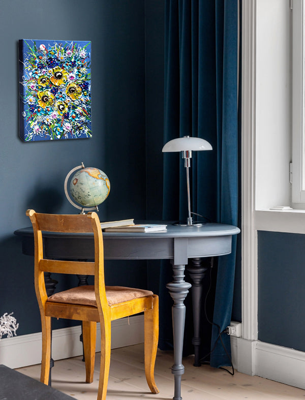 A colorful floral canvas wall art print hangs on a dark navy blue wall in a styled home office setting, featuring a wooden chair, a gray writing desk, a globe, and a table lamp.