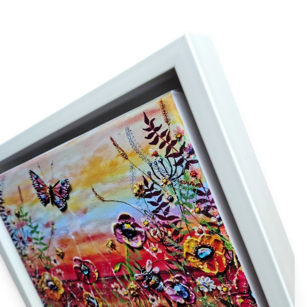 A framed floral and butterfly print on canvas shown at an angle against a white background. The artwork features a butterfly on the left and colorful flowers with yellow, orange, red, pink, purple, and blue tones against a warm sky.