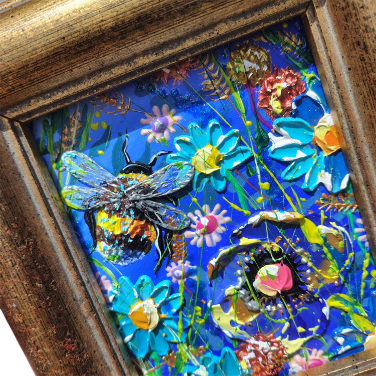 A framed colorful mixed-media artwork featuring a large textured bee over bright blue flowers and abstract splashes, set in a rustic wooden frame.