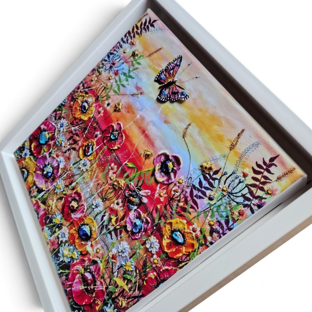A square framed canvas print featuring a colorful floral and butterfly scene, photographed at an angle against a plain white background.