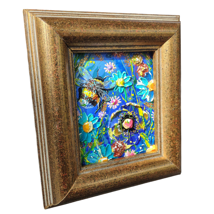A framed floral art print featuring a vibrant blue background with colorful flowers and two bumblebees, enclosed in a distressed gold-toned wooden frame.