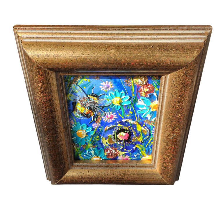 A square framed art piece featuring a colorful floral and bee illustration on a blue background, enclosed in a textured gold-toned frame against a white background.