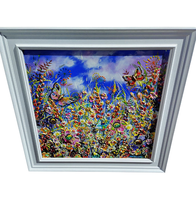 A large square framed painting depicting a vibrant field of flowers and butterflies under a blue sky, set in a white frame against a white background.